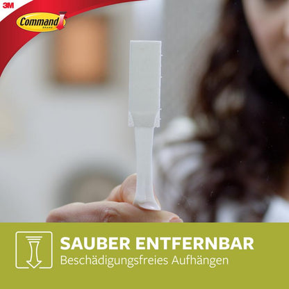 A person holds a white Narrow Command™ Picture Hanging Strip (M) 17207 from 3M Deutschland GmbH upright. The Command logo and German text about narrow picture mounting and residue-free removal are visible against a blurred background.