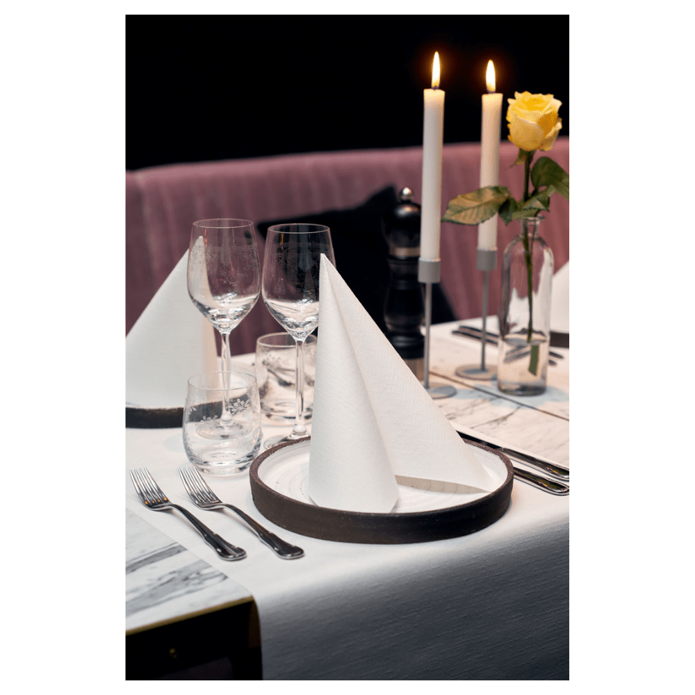 A dining table is elegantly set with two Duni Dunilin® napkins 48 x 48 cm White, folded into triangles on plates. Two large burning candles and a yellow rose in a glass vase adorn the background. Wine glasses, a water glass, and cutlery complete this sophisticated ambiance with fabric-like charm.