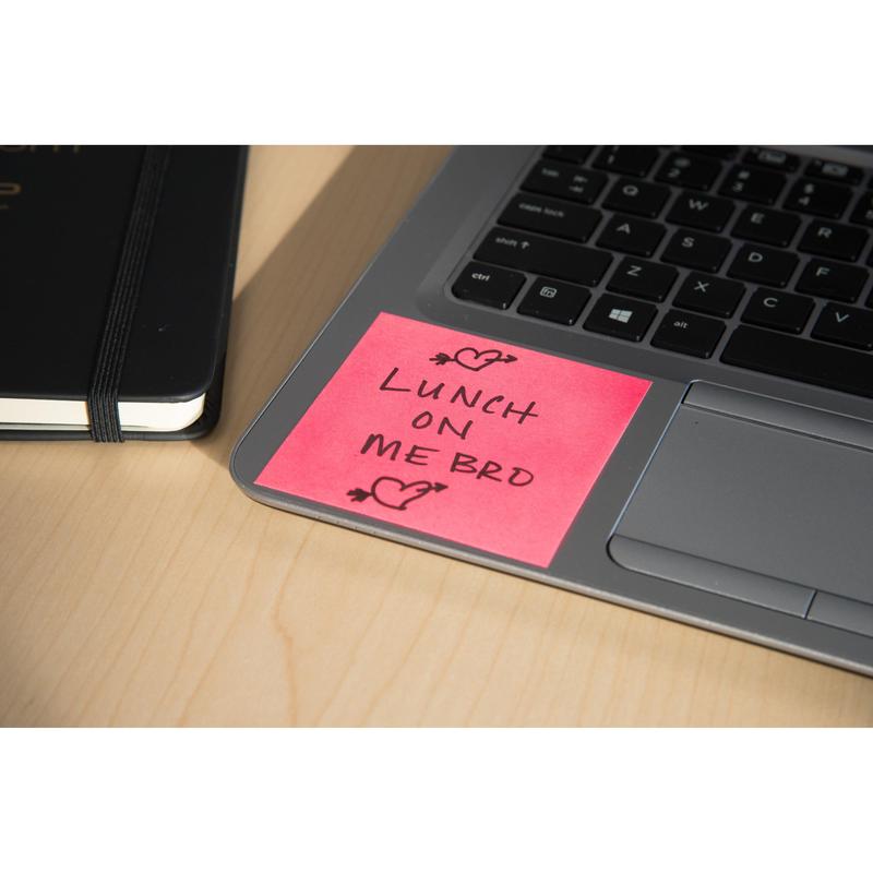 A pink Post-it® Super Sticky Z-Note (76x76mm) from 3M Deutschland GmbH with the inscription "Lunch on me bro" in black ink and two hearts lies on a laptop keyboard next to a black notebook, all on a light wooden table.
