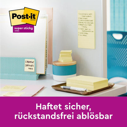 A desk with folders, pen holder, and yellow Post-it® Super Sticky Z-Notes (76 x 127 mm) from 3M Deutschland GmbH. A note sticks to the wall. German: "Sticks securely, removable without residue." Package: 12 blocks of 90 sheets, cardboard packaging.