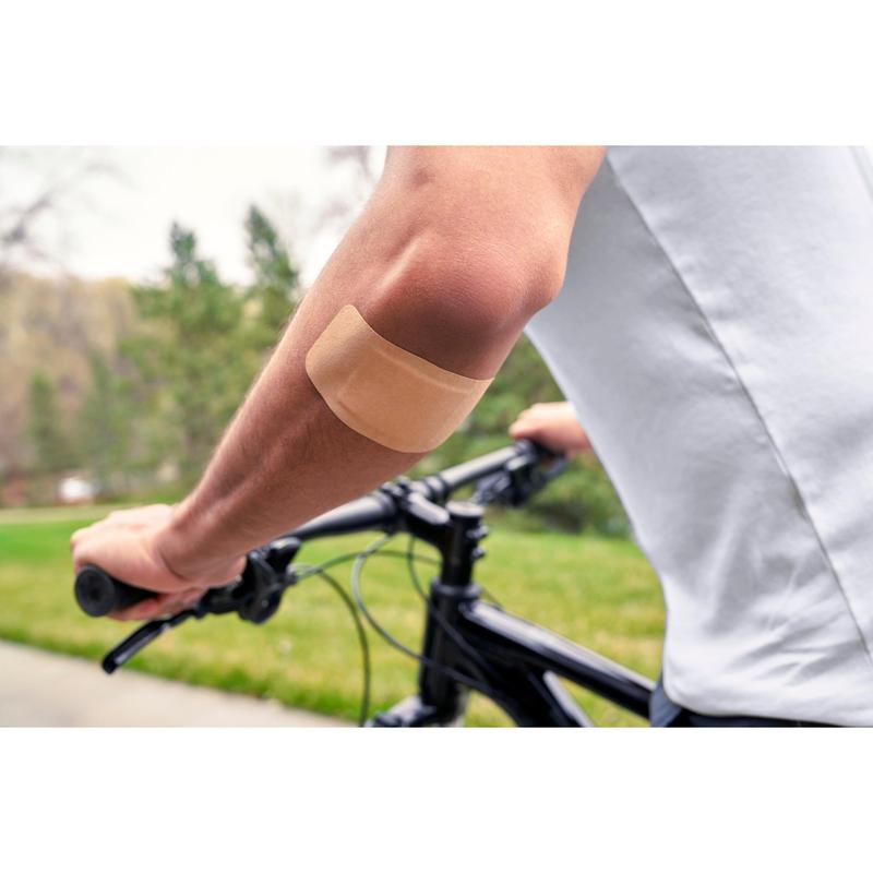 A person wearing a white shirt and cycling outdoors has a Nexcare™ Ultra Stretch MAXI Comfort Flexible Bandage (50 mm x 101 mm) from 3M Deutschland GmbH on the bare upper arm; green grass and trees are visible in the background.