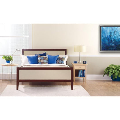 A neatly made bed with white and blue pillows stands in front of a light wall that is taped with ScotchBlue™ Painter's Tape "Multi-Surface" by 3M Deutschland GmbH. On a bedside table are a lamp, a clock, books, and a potted plant next to the bright window.