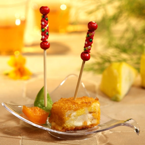 On a transparent, leaf-shaped plate lies a cube of breaded, fried food with a PAPSTAR 250 finger food pick from PAPSTAR GmbH, along with a green and an orange slice. In the slightly blurred background, a lemon slice and greens can be seen.