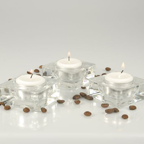Three Starpak tealights Ø 38 mm, white, with an 8-hour burn time from the folding box (100 pieces) are in square glass containers on a white surface with scattered coffee beans against a bright, simple background.