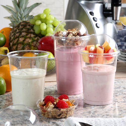 Three smoothie cups stand on a counter, two with fruit and granola in dome lids. The lids are made of Starpak 50 inserts, PET "Hurricane" round Ø 9 cm, crystal clear. In the background are fresh pineapples, grapes, and a blender.