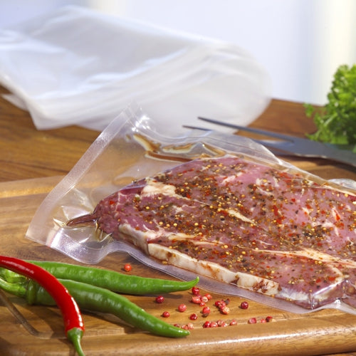 A piece of marinated raw meat, sealed in a transparent Starpak 100 vacuum bag (PA/PE), lies on a wooden cutting board, surrounded by green and red chili peppers, scattered spices, fresh herbs, and kitchen utensils.