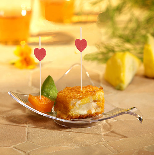 In a small glass bowl, there is a breaded, fried cube with white filling, garnished with a leaf and an orange. Two PAPSTAR 100 Fingerfood - Picks from PAPSTAR GmbH, adorned with red hearts, are included. Lemon wedges are visible in the background.