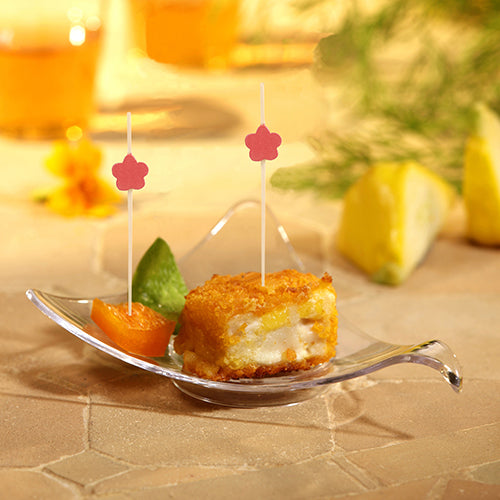 In a small glass bowl, there are breaded, layered foods and an orange slice, each with a PAPSTAR 100 Fingerfood - Pick from PAPSTAR GmbH. Next to the orange is a green garnish. Blurred fruits and drinks are visible in the background.