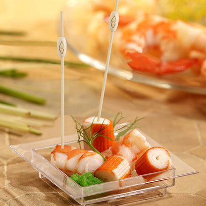 A square, transparent dish contains imitation crab, shrimp, and green roe with herbs. Two PAPSTAR 100 Fingerfood - Picks from PAPSTAR GmbH are inserted into the seafood. Blurred shrimp appear in the background.