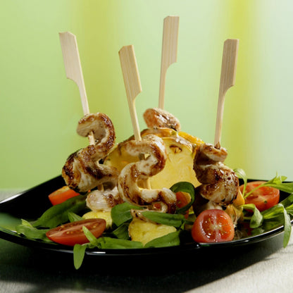 A black plate with grilled chicken skewers on greens and cherry tomatoes, arranged with PAPSTAR 250 finger food skewers "pure" by PAPSTAR GmbH around the pineapple - perfect for any party buffet. Three wooden skewers are inserted into the pineapple.