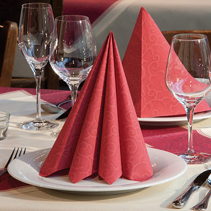 A folded napkin PAPSTAR 50 napkins "ROYAL Collection" 1/4-fold 40 x 40 cm red from PAPSTAR GmbH stands upright on a white plate, wine glasses, cutlery, and a red patterned tablecloth complete the elegant table decoration.