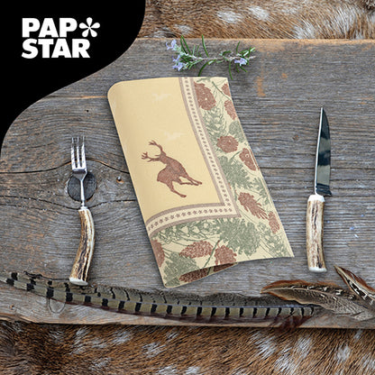A napkin from PAPSTAR GmbH ROYAL Collection (40x40 cm, 1/4-fold) with deer and pine cone motif lies on a rustic wooden table with antler-like cutlery, feathers, and herbs. In the top left corner is the PAPSTAR logo.