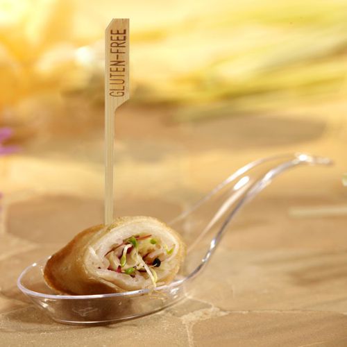 An appetizer filled with vegetables sits on a clear plastic spoon, secured with a PAPSTAR 250 finger food skewer "pure" by PAPSTAR GmbH with the inscription "GLUTEN-FREE". The gently blurred background emphasizes the attractiveness of the finger food for the party buffet.