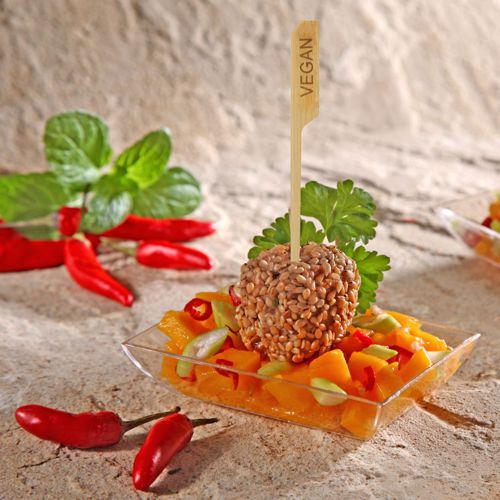 In a small clear bowl, there is a mango vegetable salad with a vegan sesame ball, served on PAPSTAR 250 finger food skewers "pure" by PAPSTAR GmbH. Garnished with parsley and mint, it is ideal for party buffets, with red chili peppers in the background.