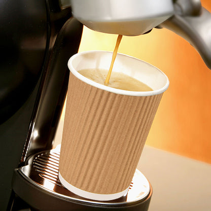 A coffee machine pours coffee into a PAPSTAR 25 drinking cup, paper "To Go" Ripple Wall from PAPSTAR GmbH, which stands on a metal drip tray. The background shows warm, blurred tones.