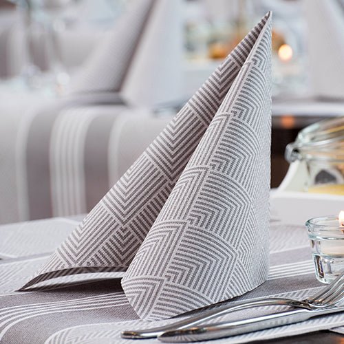 A neatly folded PAPSTAR 50 napkins "ROYAL Collection" 1/4-fold 40 x 40 cm grey by PAPSTAR GmbH stands upright on a table with fork, knife, striped tablecloth, tealight, and blurred glasses in the background.