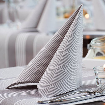 A neatly folded PAPSTAR 50 napkins "ROYAL Collection" 1/4-fold 40 x 40 cm grey by PAPSTAR GmbH stands upright on a table with fork, knife, striped tablecloth, tealight, and blurred glasses in the background.