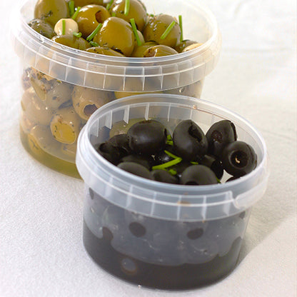 On a white surface, there are two transparent Starpak 36 deli cups with tamper-evident seals (PP, 240 ml) – one contains spiced green olives, the other sliced black olives with green garnish.