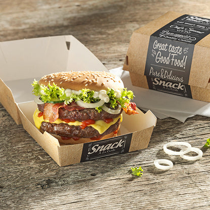 A cheeseburger with lettuce, tomato, bacon, onions, and sauce lies in an open PAPSTAR 50 burger box made of cardboard "pure" by PAPSTAR GmbH on a wooden table. Next to it, a closed box with "Good Food" print and onion rings.