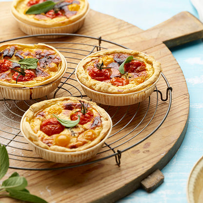 Three small vegetable quiches with cherry tomatoes and herbs lie on a round wire rack on a wooden board, surrounded by fresh herb leaves and PAPSTAR 50 baking molds "pure" brown from PAPSTAR GmbH.