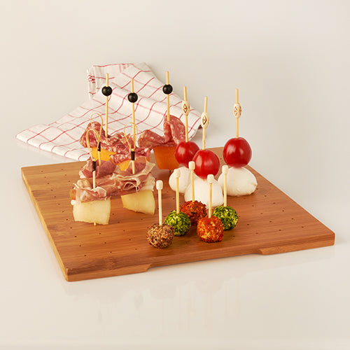 A wooden board with skewered snacks, served on the PAPSTAR holder for finger food picks, bamboo tray 25x30 cm from PAPSTAR GmbH. Cheese, sausage, tomatoes, and herb cheese balls are included; a checkered cloth can be seen in the background.