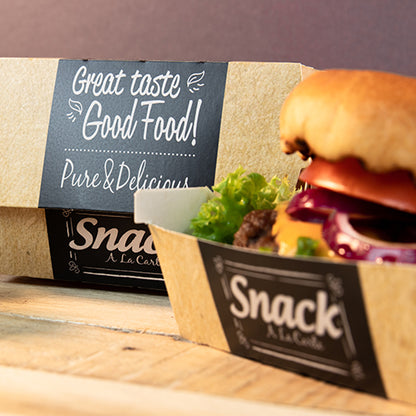 A cardboard tray with the inscription "Snack A La Carte" contains a hamburger with lettuce, tomato, and onion. In the background is a PAPSTAR 50 burger boxes, cardboard "pure" "Good Food" by PAPSTAR GmbH: Great Taste Good Food! Pure & Delicious.