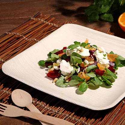 A PAPSTAR 6 plate, palm leaf “pure” square by PAPSTAR GmbH, contains a mixed green salad with beetroot, orange slices, goat cheese, and nuts. It is placed on a bamboo mat with wooden cutlery; in the background, an orange and herbs can be seen.