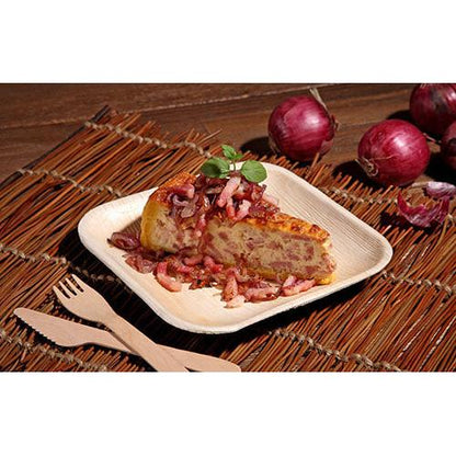 A piece of Spanish omelet with red onions is served on a square “Pure” plate from PAPSTAR GmbH from the PAPSTAR 6-piece set, accompanied by bamboo knife and fork. In the background, red onions with skin can be seen on a woven mat.