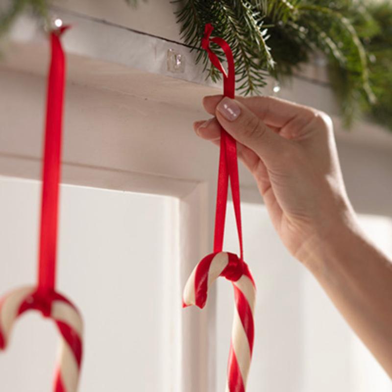 A hand hangs a candy cane with a red ribbon on a Command™ Mini Hook, transparent, with transparent strips (3M Deutschland GmbH), which is attached under a shelf decorated with evergreen branches. Another candy cane is already hanging on the hook next to it.