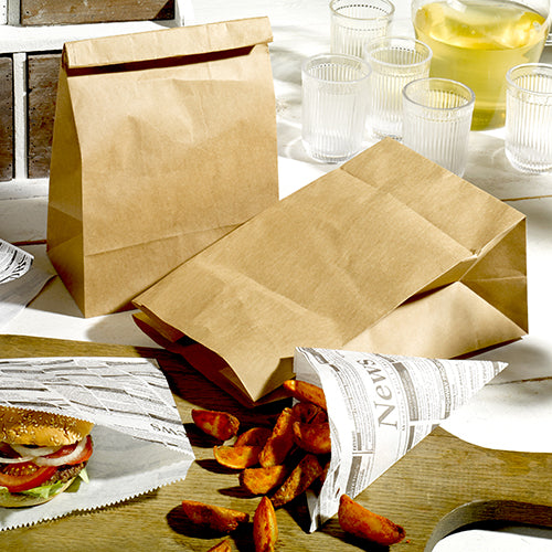 Two PAPSTAR 250 block bottom bags made of brown kraft paper from PAPSTAR GmbH, a newspaper design paper bag with potato wedges, and a burger partially wrapped in newspaper stand with four glasses and a jug on a light table.