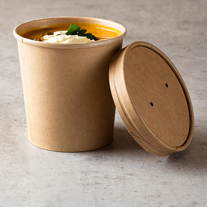A brown Starpak 25 cardboard soup cup, round, 350 ml (Ø 9.8 cm · 7 cm), is filled with soup along with cream and herbs. The matching lid is slightly offset on a light stone surface next to it.