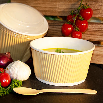 A PAPSTAR round soup cup, brown/white, Ripple Wall (13.5 cm), filled with soup, stands next to a wooden spoon on a dark surface. Nearby are vine tomatoes, garlic, onion, parsley, and another paper cup from PAPSTAR GmbH with a lid.
