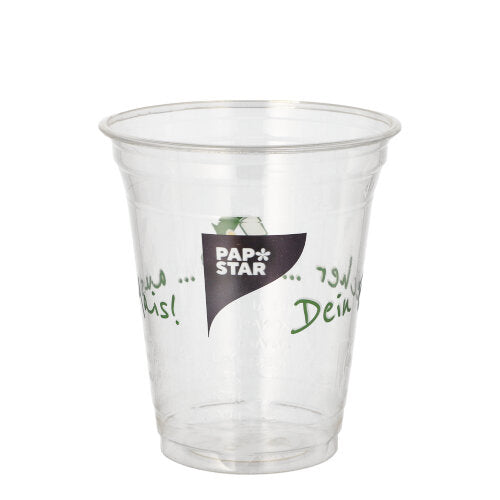 A transparent PLA cup "PAPSTAR 50 drinking cups Cup made of corn" from PAPSTAR GmbH with the logo on the front and green multilingual text on the sides is empty and presented against a plain white background.