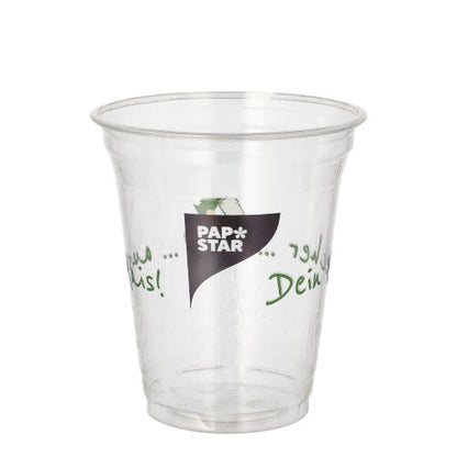 A transparent PLA cup "PAPSTAR 50 drinking cups Cup made of corn" from PAPSTAR GmbH with the logo on the front and green multilingual text on the sides is empty and presented against a plain white background.