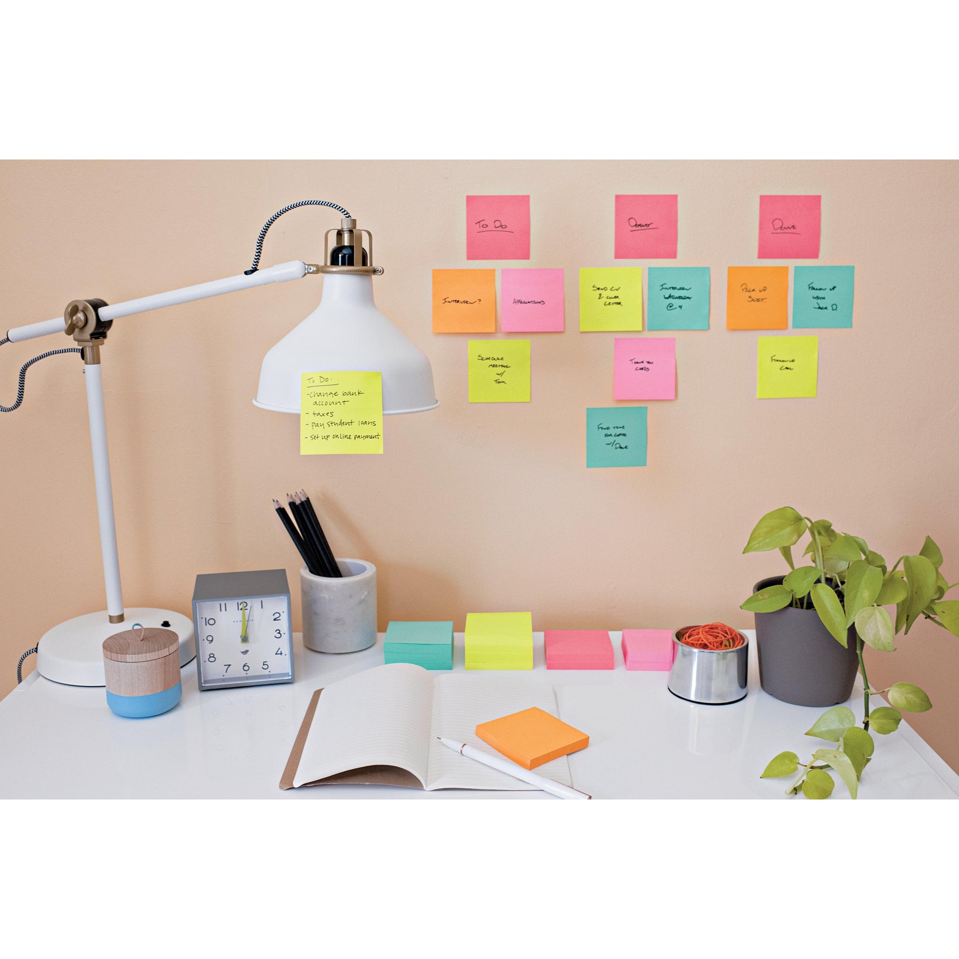Auf dem aufgeräumten Schreibtisch stehen eine Lampe, eine Uhr, eine Pflanze, Schreibwaren und ein offenes Notizbuch. Darüber zeigen bunte 3M Deutschland GmbH Post-it® Super Sticky Notes (76 mm x 127 mm, 90 Blatt/Block) handschriftliche Aufgaben und Erinnerungen an der Wand.