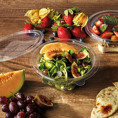 A Starpak 50 delicatessen and salad bowl with hinged lid, R-PET crystal clear round, holds a melon and fig salad on a wooden table with strawberries, grapes, bread, cherries, and fruit. Another Starpak R-PET container with fruit is nearby.