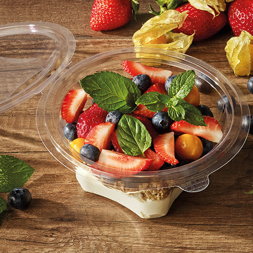 A Starpak 50 delicatessen and salad bowl with hinged lid, R-PET crystal clear round container is filled with yogurt, muesli, strawberries, blueberries, and golden berries on a wooden surface, next to fresh strawberries and mint leaves.