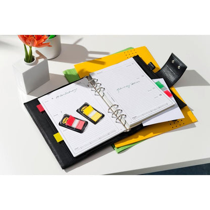 An open black leather planner with two packs of 3M Deutschland GmbH Post-it® Index Tabs (Standard, 25.4 x 43.2 mm, 50/piece) lies on a white desk with colorful folders, papers, and a small vase with an orange flower.