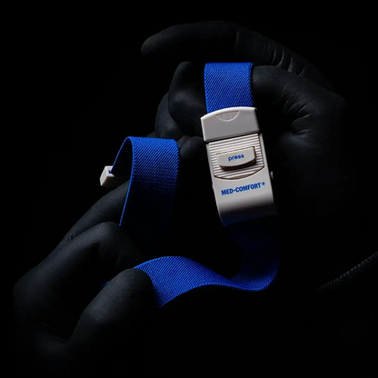 A person with black gloves holds a blue AMPri MED-COMFORT tourniquet, an indispensable instrument for medical applications. On a dark background, a white plastic buckle with a push button can be seen.
