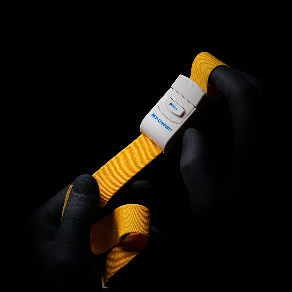 A person with black gloves holds an AMPri MED-COMFORT tourniquet, a yellow vein compressor with a white buckle, centered against a black background.
