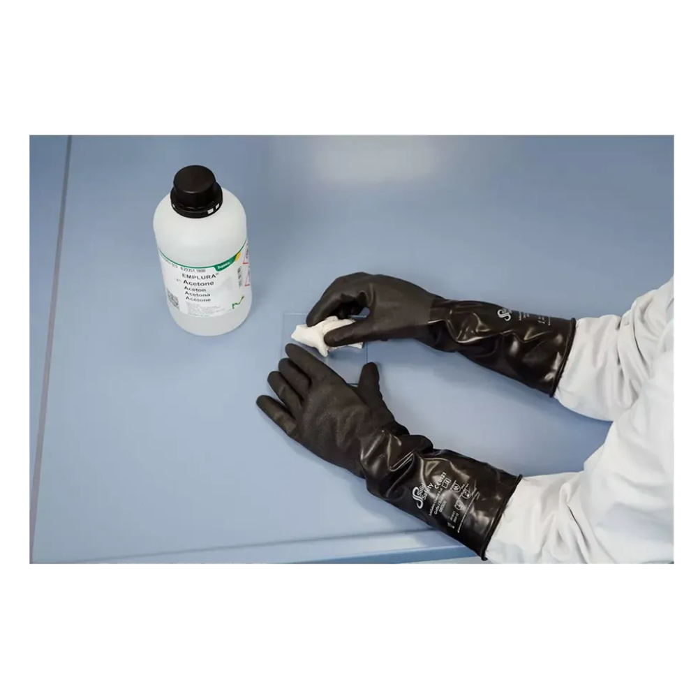 A person in a white lab coat wipes a blue surface and wears AMPri SolidSafety ChemB Full Stable Butyl Chemical Protection Gloves XXL. On the table is a bottle labeled “CAUSTIC SODA” emphasizing the importance of proper protection by AMPri Handelsgesellschaft mbH.