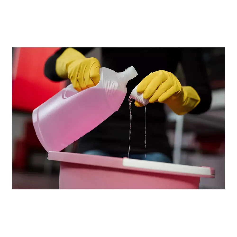 A person wearing AMPri SolidSafety ChemL chemical protection gloves by AMPri Handelsgesellschaft mbH pours liquid from a clear plastic bottle into a small cap over a pink container. The liquid, possibly an AMPri cleaning solution, stands out against the blurred background.
