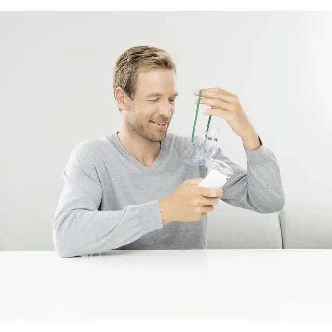 A man in a gray sweater sits at a white table and smiles while holding the Beurer Inhaler To Go IH 5. One hand holds the transparent plastic nasal mask while the other skillfully attaches the green strap, showcasing the advanced oscillating membrane technology. The background is simple and bright.