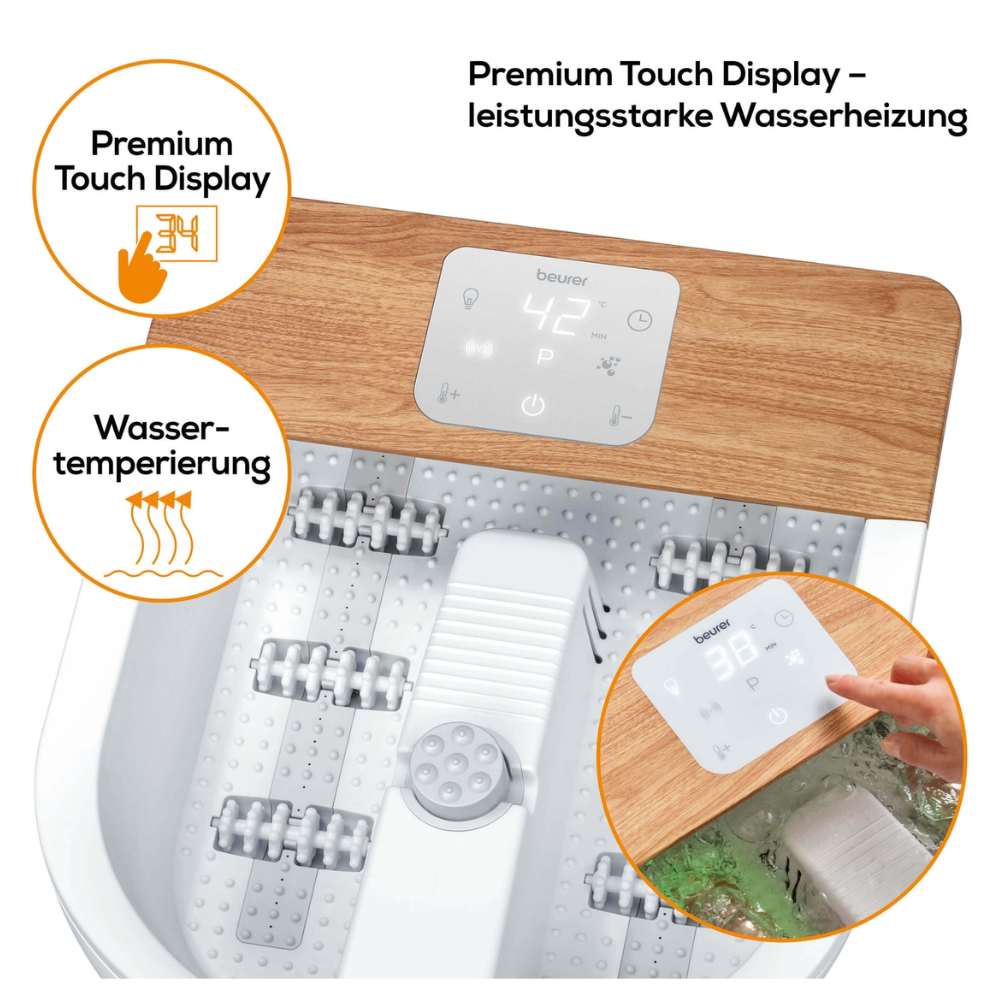 A foot spa with the brand Beurer FB 65 Wellness foot spa from Beurer GmbH has a white base and a wooden plate, with a digital touch display showing a temperature of 42 °C. The labels read "Water temperature" and "Premium Touch Display." A hand adjusts the display and massage rollers for vibration and bubble massage are visible.