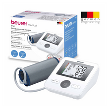 The image shows a Beurer upper arm blood pressure monitor BM 27 | Package (1 piece) from Beurer GmbH with its packaging box. The device features a digital display, arrhythmia detection, and an attached cuff. The packaging highlights important features and characteristics, including a “German Engineering” logo in the upper right corner.