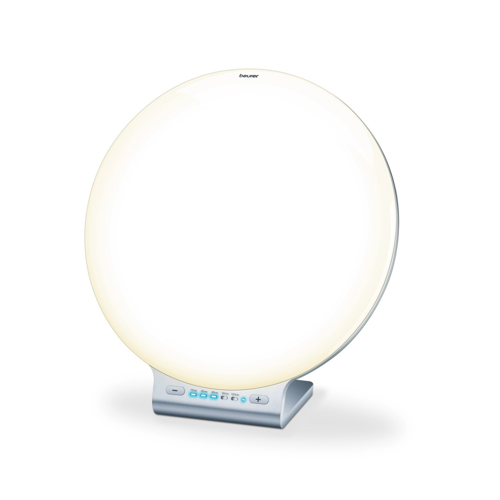 A round, white Beurer daylight lamp TL 100 from Beurer GmbH on a silver stand with a small control panel at the bottom with buttons and a digital display. The lamp emits a soft, even light, perfect as mood lighting, with the brand name "Beurer" printed at the top center.