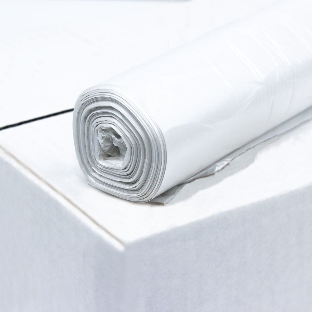 A roll of DEISS HDPE garbage bags 20 liters, 59906, white from EMIL DEISS KG (GmbH + Co.) lies on the edge of a white surface made from recycled material. The bags are neatly rolled and have multiple layers. The plain white background emphasizes the texture and high film elasticity of the plastic.
