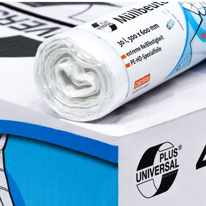 A roll of DEISS Universal Plus® garbage bags, 30 liters, from EMIL DEISS KG (GmbH + Co.) with blue and orange lettering lies on a carton. The text includes "garbage bags" as well as information about the HDPE special film, dimensions, and material. In the background, another roll is partially visible.