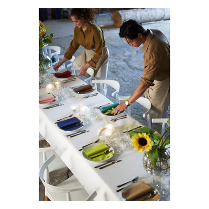 Two people set a long table with a white tablecloth from Duni GmbH Dunicel tablecloth rolls, plates, glasses, and colorful napkins in green, yellow, and pink. A vase with sunflowers serves as table decoration. The interior is equipped with atmospheric lighting.