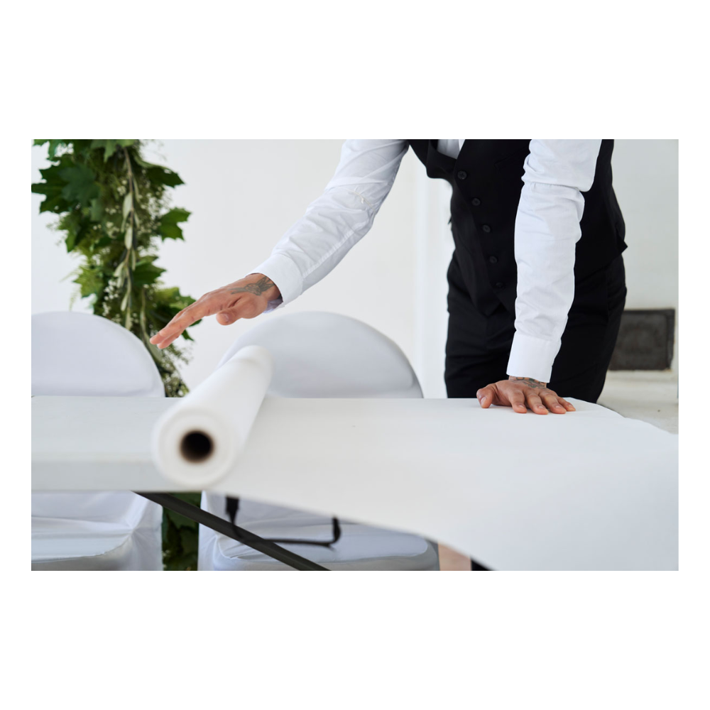 A person in evening attire, with a white shirt and black vest, rolls out a Dunicel tablecloth roll (1.18 m x 40 m) from Duni GmbH over a table; some green is visible against the plain background.